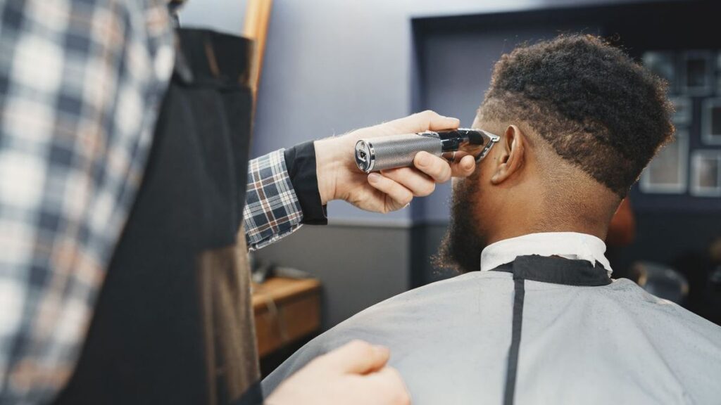 Barber in a plaid shirt trimming the side of a bearded client's hair with a cordless trimmer